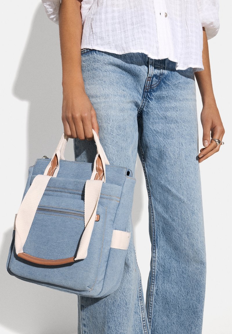 Blaue Denim-Tote- Tasche mit hellen Canvas-Tragegriffen, ausgestattet mit einer vorderen Reißverschlusstasche und einem Akzent aus braunem Leder. Robuste, quadratische Form, lässiges Design.