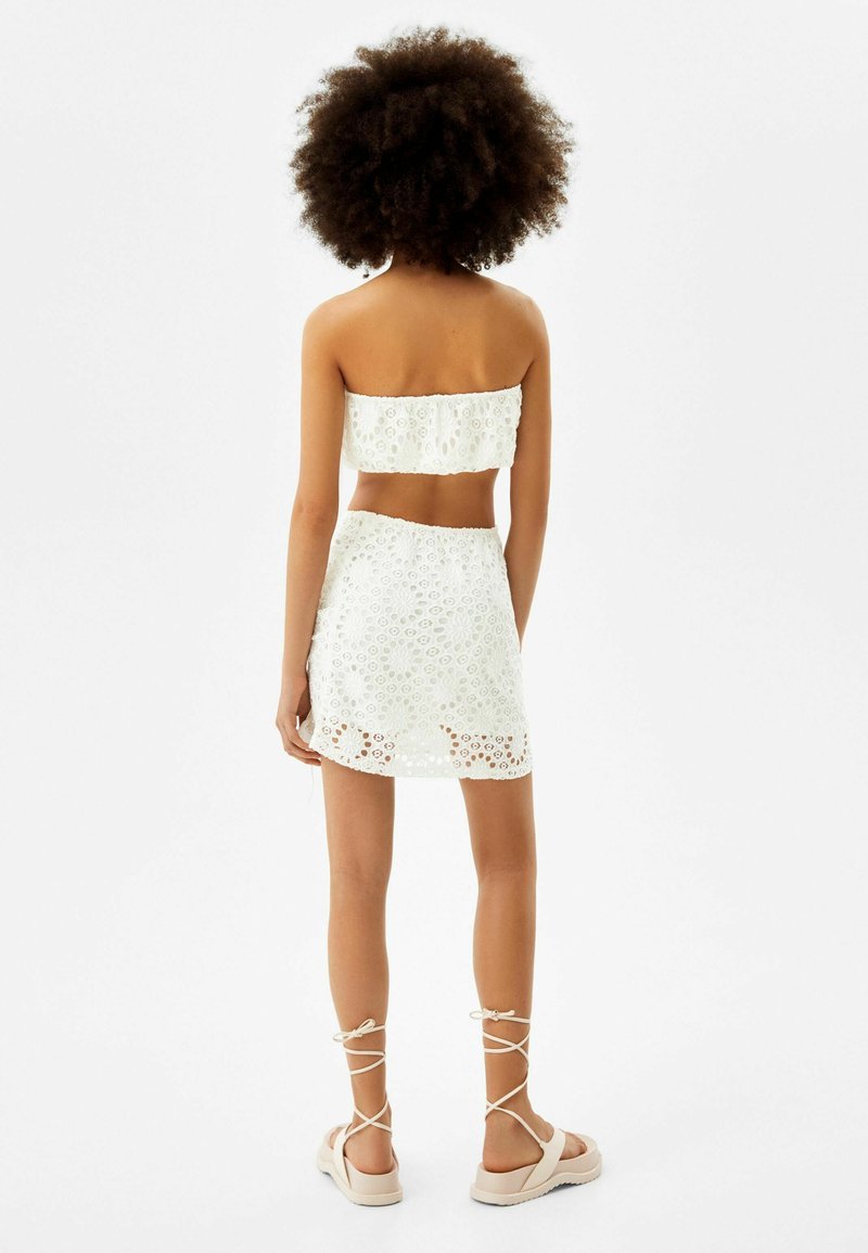Weißes Häkel-Zweiteiler-Set mit einem Bandeau-Top und einem Minirock mit Blumenmustern. Kombiniert mit sandfarbenen Riemchensandalen.