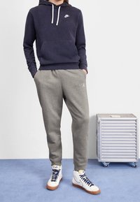 Granatowa bluza z kapturem z białym logo, szare spodnie dresowe z detalem logo oraz białe wysokie trampki z niebieskimi sznurówkami i brązowymi podeszwami.