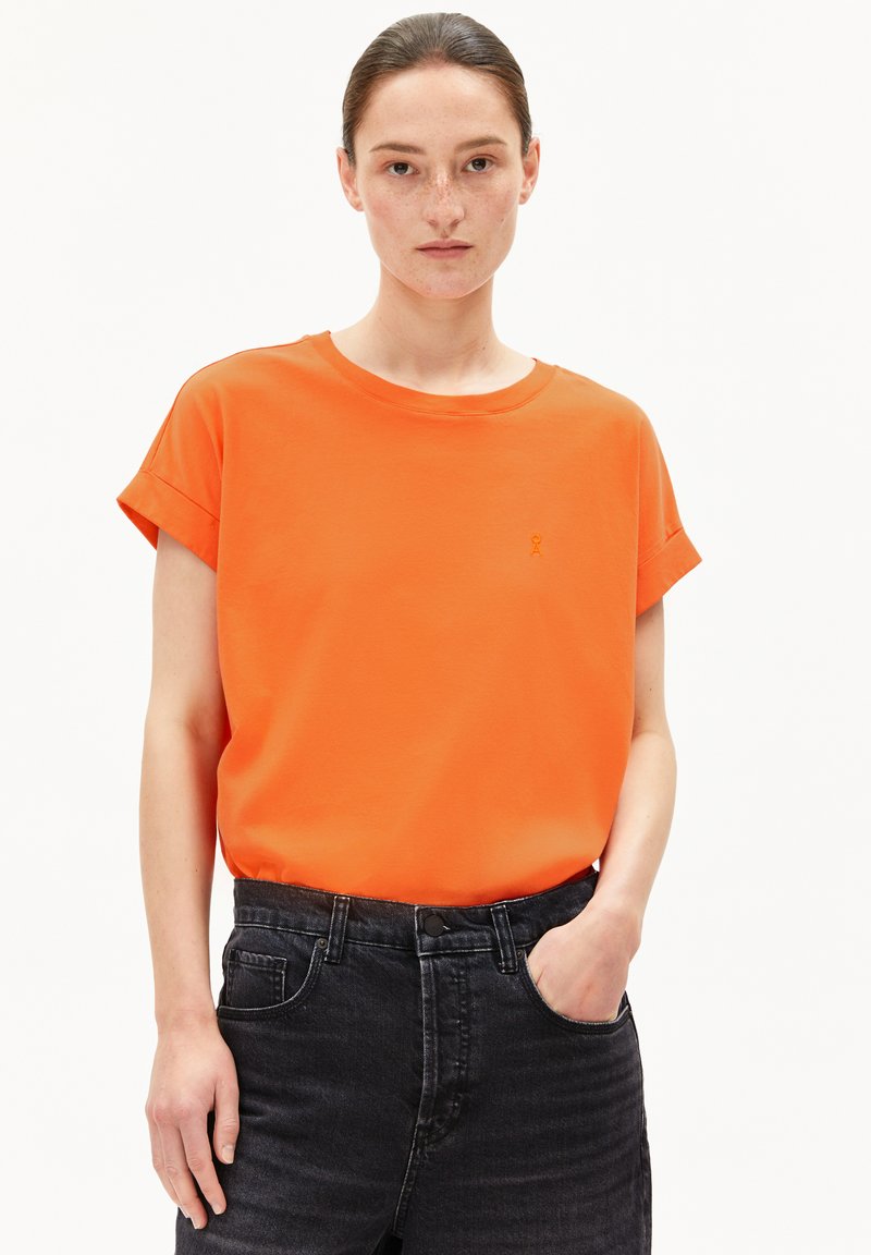 Helles orange T-Shirt mit kurzen Ärmeln, rundem Ausschnitt und umgeschlagenen Ärmeln, mit einem kleinen gestickten Logo auf der linken Brust. Getragen mit schwarzen Jeans.