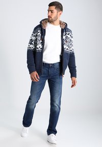 Marine strikket cardigan med hvite snøfnuggmønstre, plysjforet hette, frontglidelås, kombinert med blå jeans og hvite sko.