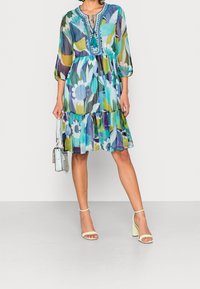 Šaty s květinovým potiskem, se třičtvrtinovými rukávy, modro-zelenými odstíny, vrstveným spodním lemem, výstřihem ve tvaru klíčové dírky a složitým detailem na výstřihu.
