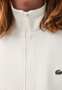 Giacca bianca con zip realizzata in un tessuto testurizzato e un piccolo logo di coccodrillo verde sul lato sinistro del petto. Design con colletto alto.