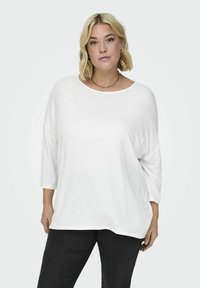 Camisa blanca de gran tamaño, de manga larga, con un escote ancho y hombros caídos, fabricada en un tejido suave. Combinada con pantalones negros ajustados.