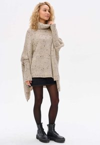 Beżowy, oversize'owy sweter dzianinowy z wysokim kołnierzem, teksturowanym wzorem i podwiniętymi rękawami, w połączeniu z czarną spódnicą i lace-up'owymi butami bojowymi.