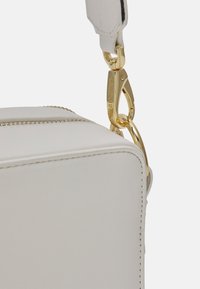 Väskan i vitt läder har en rektangulär form. Den har ett guldmetallspänne och dragkedja, samt söm-detaljer längs kanterna.