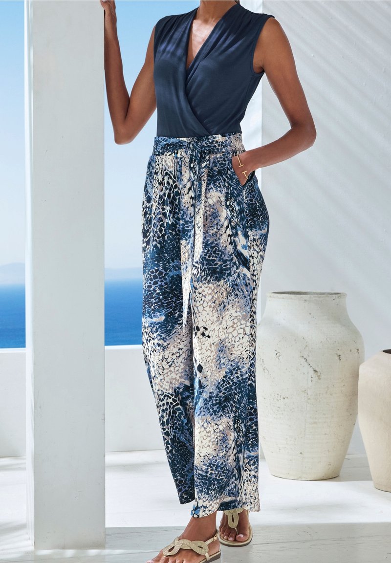 Femme portant un top sans manches bleu marine rentré dans un pantalon large taille haute avec un motif abstrait bleu et beige, debout près d'une colonne blanche.