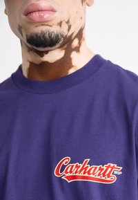 Purple cotton t-shirt featuring "Carhartt WIP" logo in red script on the front. Round neckline and short sleeves. Smooth texture.