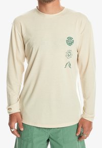 Langärmliges Cremehemd mit grünen Designs aus Spiralen und Friedenssymbolen auf der Brust und dem Ärmel; aus weichem, leichtem Stoff gefertigt.