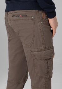 Brown cargo pants made of cotton with multi-functional pockets, contrasting stitching, and a decorative label on the back pocket.