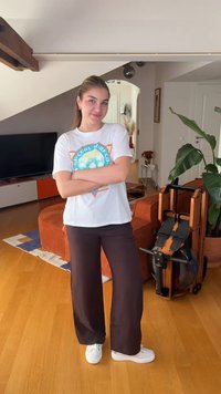 Νεαρή γυναίκα με καθημερινά ρούχα στέκεται με τα χέρια σταυρωμένα σε ένα σαλόνι που διαθέτει τηλεόραση, πορτοκαλί καναπέ, μηχάνημα κωπηλασίας και μεγάλο πράσινο φυτό.