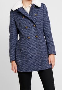 Kvinna i en blå texturerad dubbelknäppt kappa med guldbitar och en vit fuskpälskrage, kombinerad med svarta byxor.