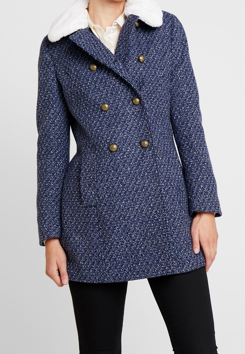 Kvinna i en blå texturerad dubbelknäppt kappa med guldbitar och en vit fuskpälskrage, kombinerad med svarta byxor.