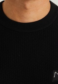 Sweater nero a maglia a coste con scollo rotondo e una piccola etichetta logata sul lato inferiore. La texture è visibile, con un tessuto strutturato.