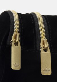 Svart sammetspåse med två guldfärgende dragkedjor med logotypade draghandtag. Närbild som framhäver den släta texturen och blanka detaljer i metall.