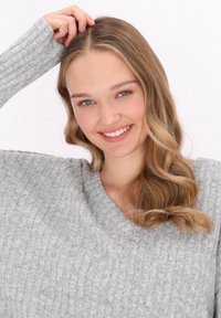 Világosszürke, bordázott pulóver V-nyakkivágással, amely lágy textúrát mutat. A modellen hosszú, hullámos haj és ragyogó mosoly található.