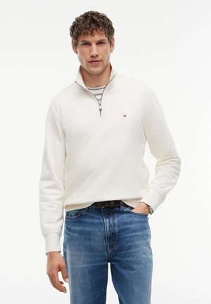 Hombre con suéter blanco con cremallera hasta el pecho sobre camisa de rayas, con la mano en el bolsillo, combinado con vaqueros azules y un reloj en la muñeca izquierda.