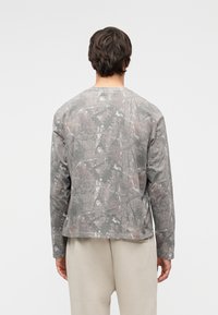 Mees, kes kannab halli kamuflaažimustriga pika varrukaga särki ja helebeezhiseid pükse, seisab valge tausta ees seljaga vaataja poole.