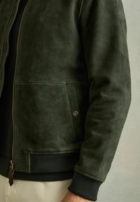 Jachetă din suede verde închis, cu închidere prin fermoar, buzunar lateral și mansete ribbate. Textură netedă cu detalii de cusătură minime.