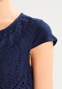 Top azul marino con intrincados bordados troquelados en la parte frontal, mangas cortas y una textura suave en los hombros.
