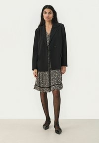 Zwarte blazer met een enkele knoop, gedragen over een getextureerde zwart-witte jurk met een rimpelonderrand. Matte zwarte puntige flats.