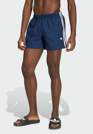 Mann trägt dunkelblaue Adidas-Sportshorts mit weißen Streifen und schwarze Adidas Slide-Sandalen und steht vor einem schlichten Hintergrund.