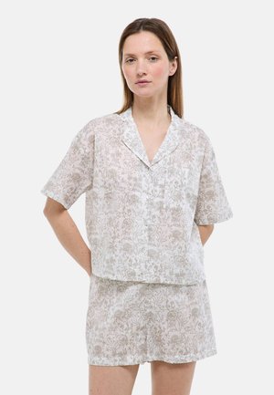 Femme portant un ensemble assorti chemise à manches courtes à boutons et short à fleurs de couleur claire, debout les mains derrière le dos sur un fond uni.