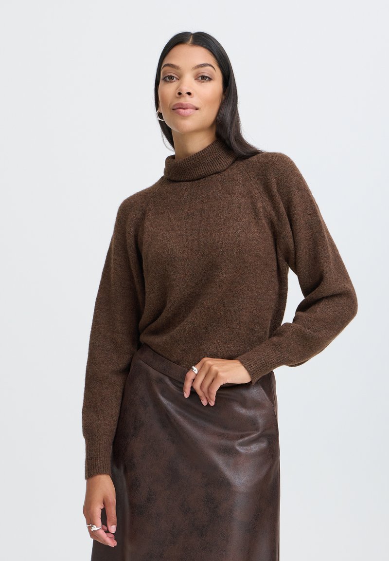 Femme aux longs cheveux foncés porte un pull à col roulé marron et une jupe en cuir marron foncé, posant avec une main sur la taille.