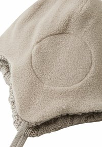 Beige fleecemuts met een gestructureerde afwerking, voorzien van een ronde patch en gebreide rand aan de onderkant. Inclusief trekkoord voor aanpassing.