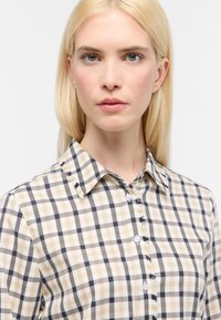 Camicia a scacchi beige e blu navy con colletto button-down, caratterizzata da bottoni bianchi e bordi sfrangiati lungo il colletto e la patta.
