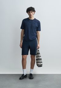 Μπλε ναυτικό ριγέ μπλουζάκι και μπλε σορτς, κατασκευασμένα από ελαφρύ ύφασμα. Κρατάει μια τσάντα tote με ασπρόμαυρο σχέδιο ζιγκ ζαγκ. Μαύρα μοκασίνια και άσπρες κάλτσες.