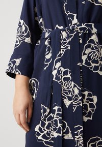 Mörkblå morgonrock med stora vita blommönster, knutet midjebälte och trekvartslånga ärmar i ett mjukt, texturerat tyg.