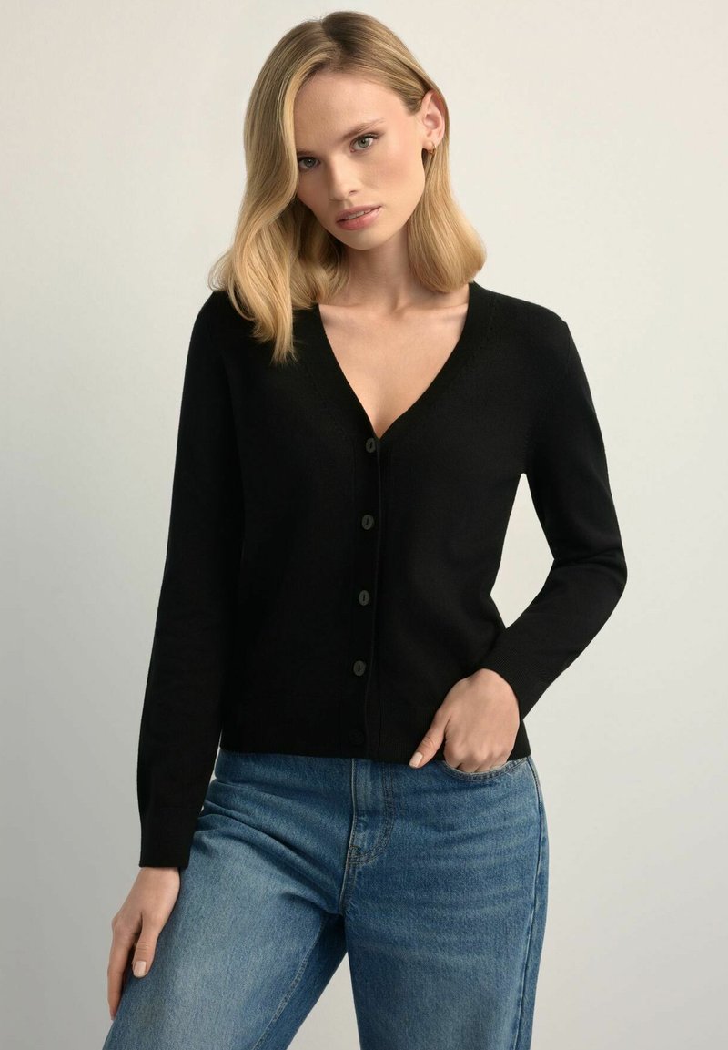 Czarny kardigan z dekoltem w serek, z zapięciem na guziki z przodu i długimi rękawami. Noszony na jasnoniebieskich dżinsach, o gładkiej fakturze.
