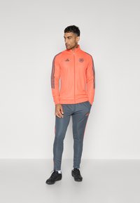 Orangene Sportjacke mit Reißverschluss und drei schwarzen Streifen an den Ärmeln, kombiniert mit grauen Hosen mit orangefarbenen seitlichen Streifen und schwarzen Sneakern.