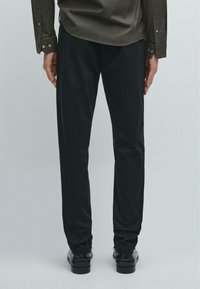 Černé ušité kalhoty s hladkou texturou, rovný střih a jemné švy, doplněné tmavými botami a zelenou košilí.