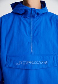 Kék kapucnis dzseki félig cipzárral és "JORDAN" feliratú logóval a zseb fedelén, viselve egy személy által, csak a felső test látható.