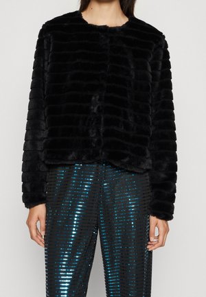 Kobieta nosząca czarną, teksturowaną kurtkę z futra ekologicznego i czarne spodnie z niebieskimi, cekinowymi pionowymi paskami.