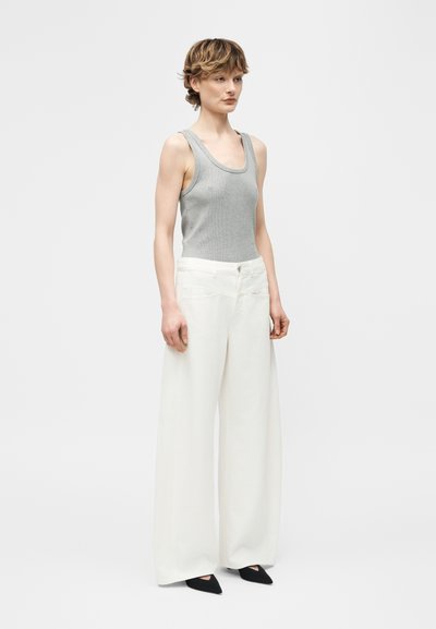 Personne portant un débardeur gris sans manches, un pantalon blanc taille haute à jambes larges, et des chaussures noires à bouts pointus, debout devant un fond uni.