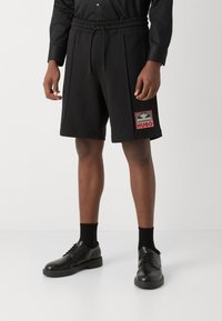 Shorts noirs avec une ceinture élastique, des poches latérales et un patch logo rouge et gris. Portés avec des chaussures en cuir noir et des chaussettes.