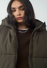 Olivgrüne Steppjacke mit einer passgenauen Kapuze, mit Reißverschluss und subtiler glänzender Oberfläche, über einem dunkelbraunen Strickpullover und einer silbernen Kette.