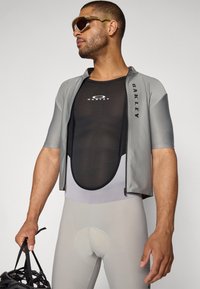 Tenue de cyclisme athlétique comprenant un haut à manches courtes gris clair, une couche en maille noire et un legging rembourré gris clair. Les accessoires comprennent des lunettes de soleil.