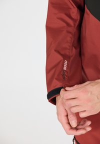 Röd vattentät jacka med svarta accenter och mjuka fleece-muddar. Har "WP 8000"-logotypen nära ärmen, vilket visar en smidig och stilren design.
