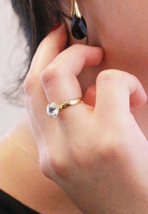 Gouden ring met een grote, ronde, heldere steen en een gladde band; zwarte druppelvormige oorbel met een gouden accent; hand die het gezicht aanraakt.
