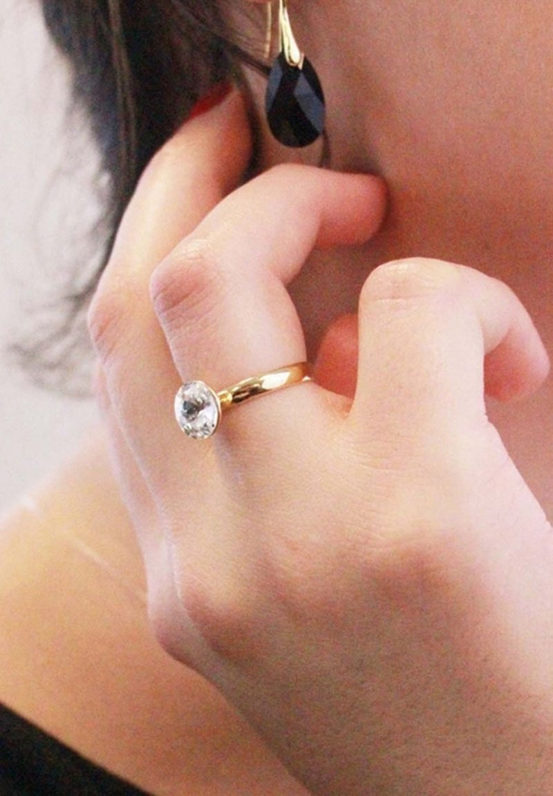 Anello d'oro con una grande pietra rotonda e trasparente e una banda liscia; orecchino a goccia nera con un accento dorato; mano che tocca il viso.