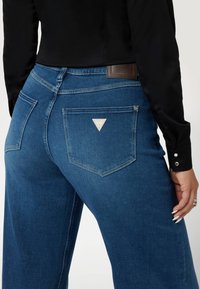 Aukštai juosmeniški mėlyni džinsai su lygiu džinsiniu audiniu, trikampiu logotipu ant nugaros ir šoninėmis kišenėmis.