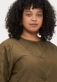 Ελαιόχρωμο επενδυμένο μπουφάν με κουμπιά μπροστά, που διαθέτει στρογγυλεμένες ραφές και λεία, γυαλιστερή υφή.