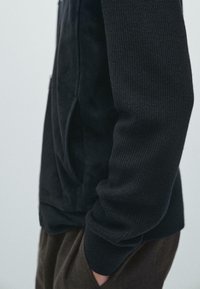 Felpa nera con cappuccio realizzata in tessuto a maglia testurizzata con accenti lisci, dotata di una tasca laterale. La manica è a costine, aggiungendo dimensione al design.