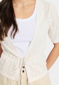 Gai beige adīta džemperis ar atvērtu priekšu, īsām piedurknēm un auklu jostasvietā. Iezīmējas ar zigzaga rakstu un ribota apdare.