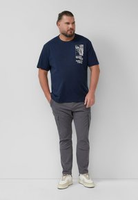 Man met een baard en achterop gebonden haar staat met handen in de zakken, gekleed in een marineblauw grafisch t-shirt, grijze cargobroek en witte sneakers.