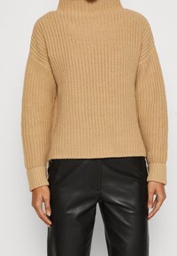 Suéter beige de punto con cuello alto, mangas anchas y textura acanalada. Combinado con pantalones negros de cuero sintético, mostrando un ajuste relajado.